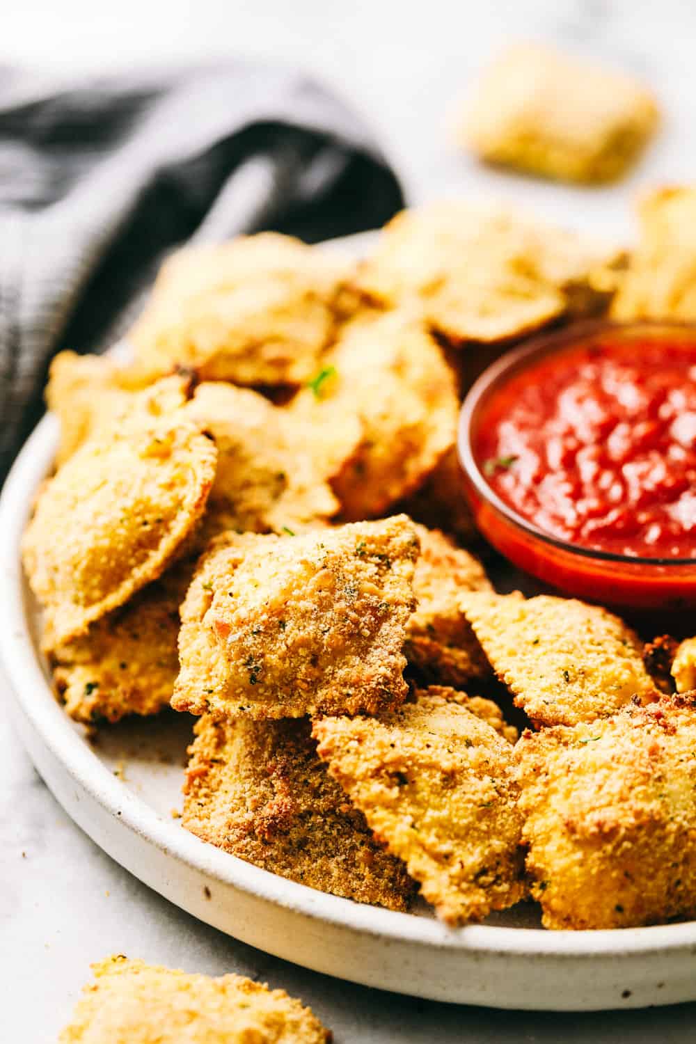 Crispy Baked Parmesan Ravioli are crunchy, crispy and filled with cheese and are perfect for dipping! These little bites are so addicting and good and will become a family favorite! Crispy Baked Parmesan Ravioli stacked on a plate with sauce.