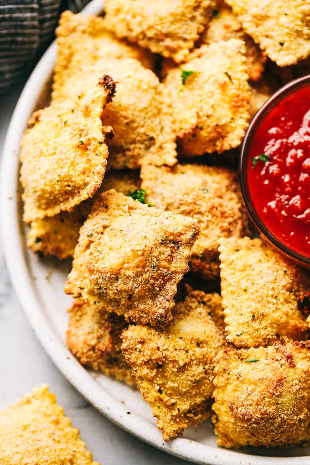 Crispy Baked Parmesan Ravioli are crunchy, crispy and filled with cheese and are perfect for dipping! These little bites are so addicting and good and will become a family favorite! Crispy Baked Parmesan Ravioli stacked on a plate with sauce.