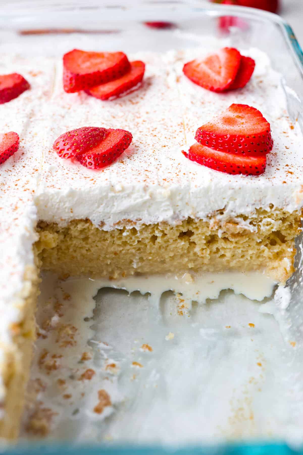 This tender Tres Leches cake is drenched in a three-milk mixture and is topped with fluffy whipped cream, cinnamon, or fresh fruit for a truly authentic Mexican dessert. Close up shot of tres leches cake with a piece taken out of it so you can see the milk mixture seeping out of the bottom of the cake.