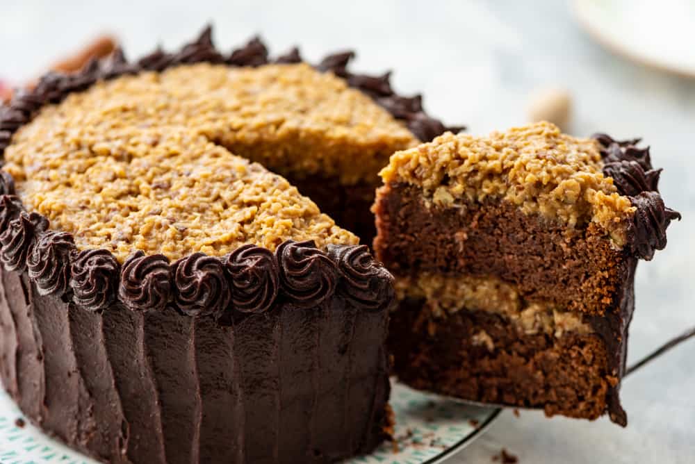 Homemade German Chocolate Cake, with two layers of scratch made cake, a sticky coconut pecan topping, and fudge frosting is everyone's favorite cake! Cutting a slice of Homemade German Chocolate Cake.