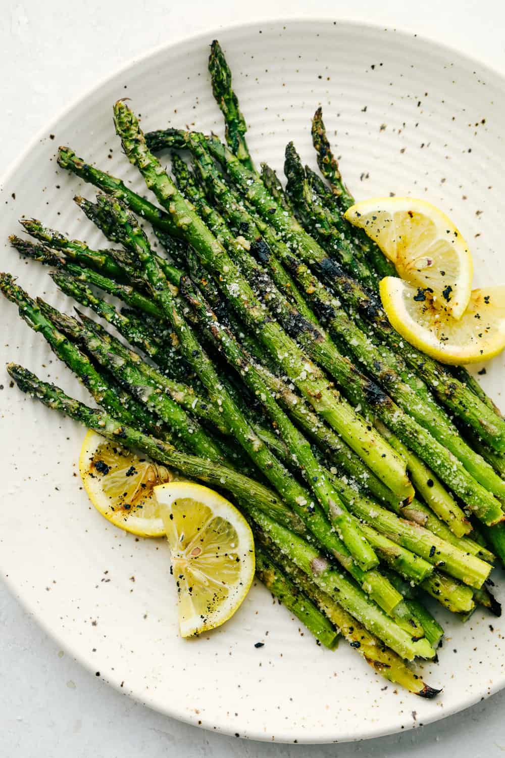 Fresh Asparagusย that is grilled gives you char marks and a smoky flavor that is amazing! Within minutes you have tender asparagus coated in parmesan and garlic that will become a family favorite! Grilled Asparagus on a white plate garnished with lemon wedges.