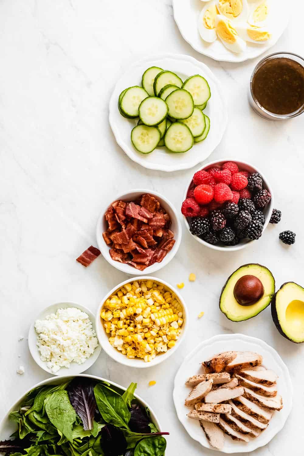 Summer Cobb Salad is filled with fresh summer fruits and veggies. It's loaded with grilled chicken, bacon, hard boiled eggs, avocado, goat cheese, berries, cucumber, grilled corn and tossed in a simple balsamic lime vinegar dressing. Every bite is refreshing, bright, and summery! Summer cobb salad ingredients in separate bowls.