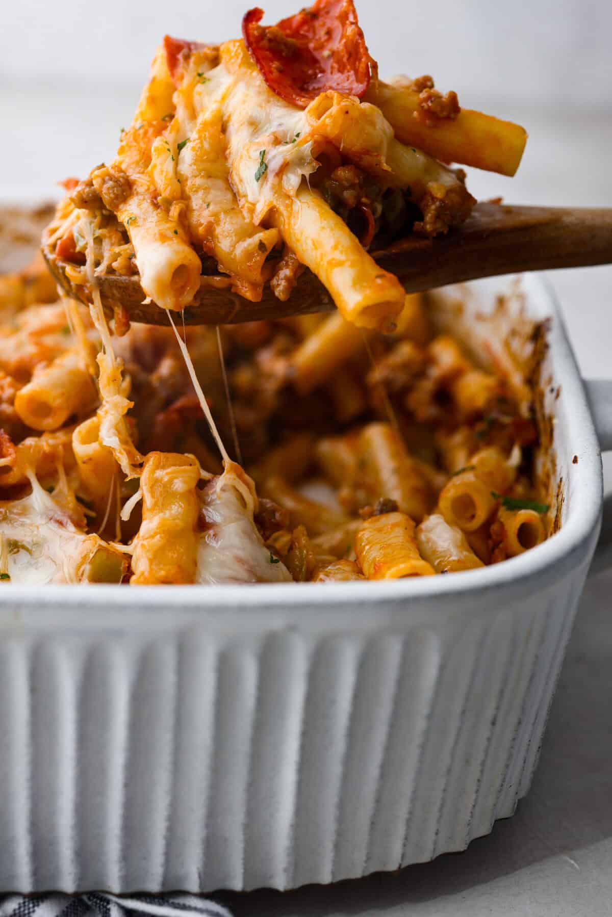 This cheesy baked ziti is baked to perfection with al dente noodles, spicy pepperoni, flavorful sausage, and smothered in melty cheese. It comes together quickly and easily and can be made ahead of time for busy weeknights! Side shot of someone scooping a portion of cheesy baked ziti out of a baking dish.