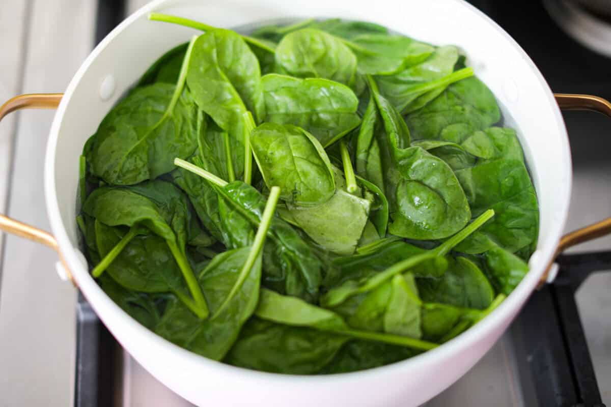 Creamed Spinach is a classic and delicious side dish! It is loaded with fresh spinach and has the creamiest garlic parmesan sauce. Serve it alongside any meal! First photo of the spinach blanching in a pot.