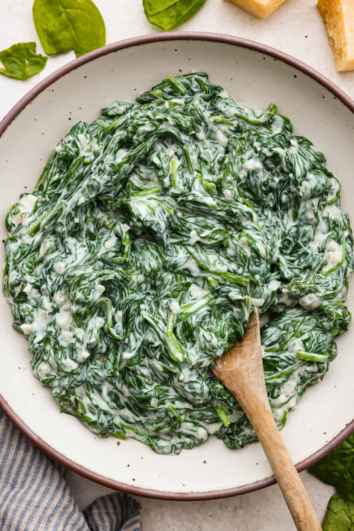 Creamed Spinach is a classic and delicious side dish! It is loaded with fresh spinach and has the creamiest garlic parmesan sauce. Serve it alongside any meal! Top view of creamed spinach in a serving bowl with a wood spoon.