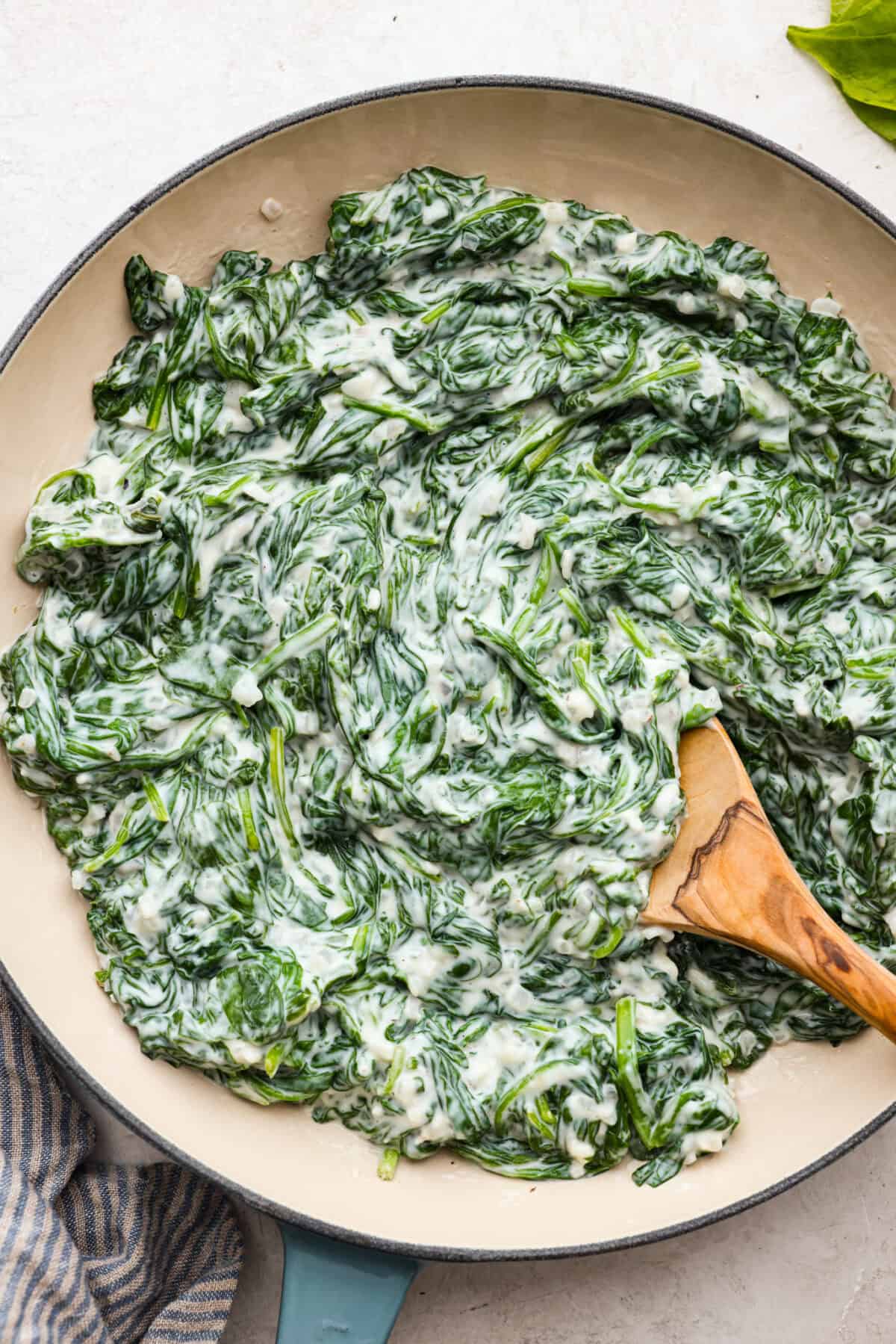 Creamed Spinach is a classic and delicious side dish! It is loaded with fresh spinach and has the creamiest garlic parmesan sauce. Serve it alongside any meal! Top close view of creamed spinach in a serving bowl with a wood spoon.