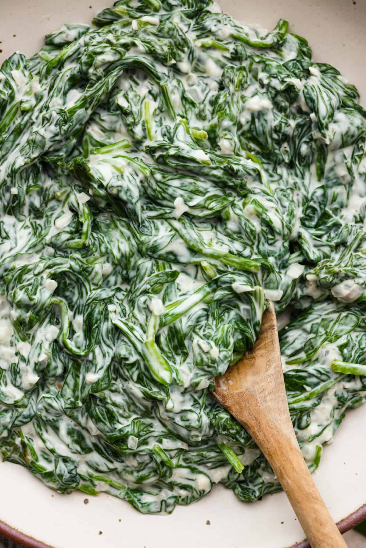 Creamed Spinach is a classic and delicious side dish! It is loaded with fresh spinach and has the creamiest garlic parmesan sauce. Serve it alongside any meal! Close view of creamed spinach in a serving bowl with a wood spoon.