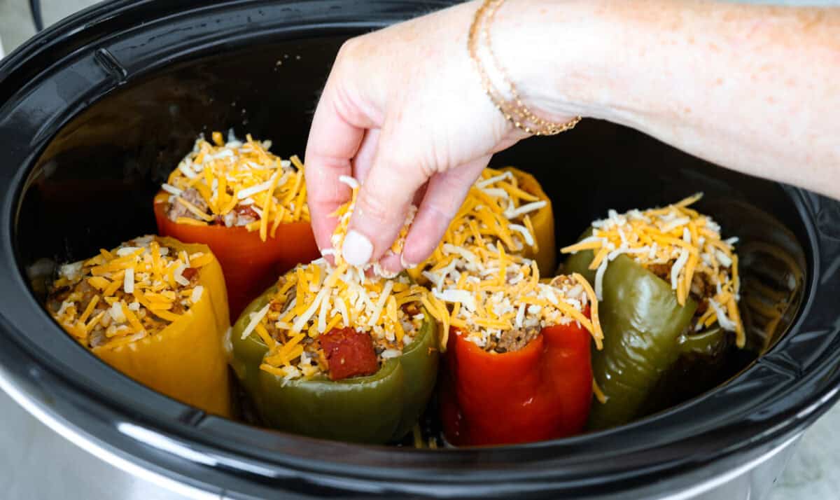 Slow cooker stuffed bell peppers are packed with ground beef, wild rice, diced tomatoes, soft black beans, and melty cheese. They cook to perfection in the slow cooker and make a healthy, delicious dinner your whole family will love! Angle shot of someone sprinkling cheese on top of bell peppers in crock pot.