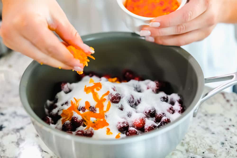 This cranberry sauce is the perfect mixture of sweet and tart! It's made with fresh cranberries and topped with orange zest to really set it apart! Cranberries in a pan with sugar and adding orange zest overtop.