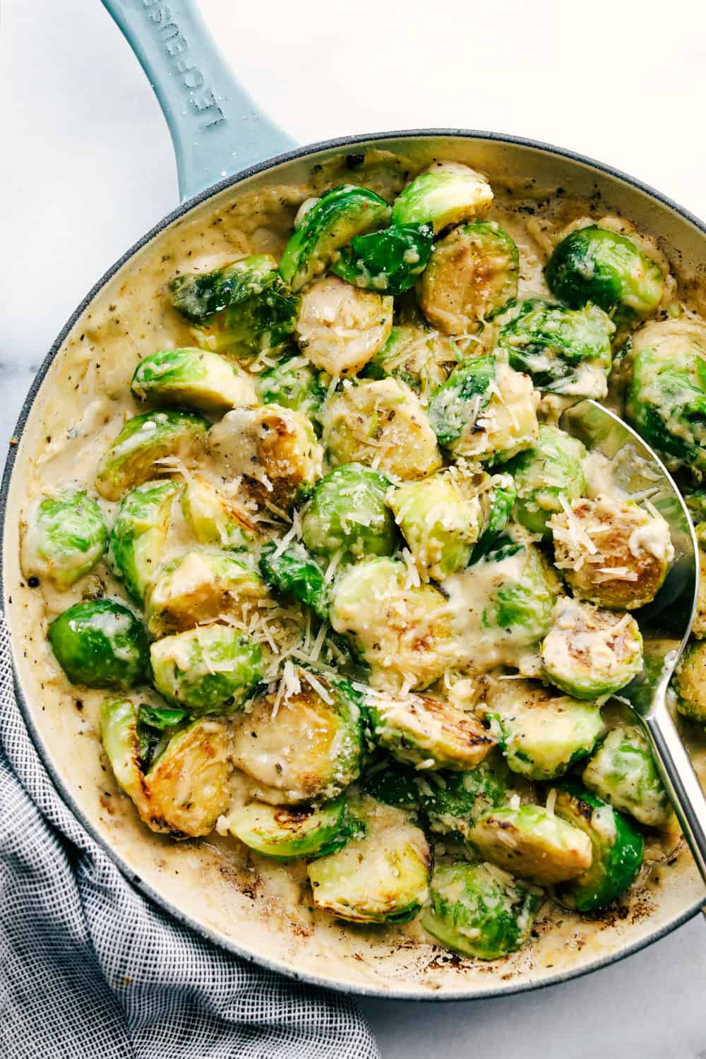 Creamy parmesan garlic Brussels sprouts sauteed and smothered in a rich and cheesy parmesan garlic sauce. Elevate your Brussels sprouts with this easy and delicious side dish! Top view of creamy parmesan garlic Brussels sprouts in a skillet with a silver serving spoon.