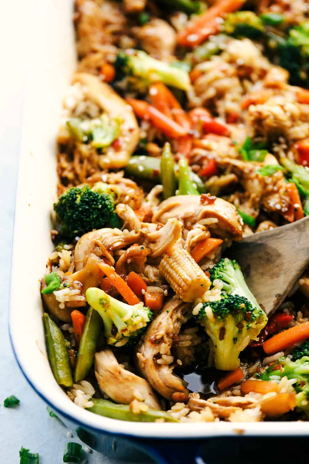 Easy Teriyaki Chicken Casserole isย a simple casserole made with homemade teriyaki sauce, vegetable stir fry and rice that's all baked to perfection. A savory dish that everyone loves!ย Scooping up chicken, vegetables, and rice with a wooden spoon.