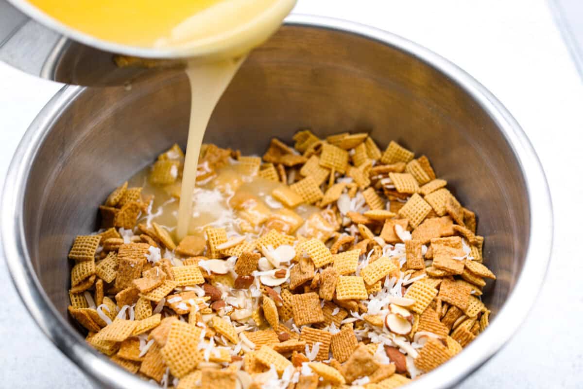 Ooey Gooey Chex Mix is the perfect sweet, salty, and crunchy blend. It has a mixture of cereal, sliced almonds, and shredded coconut, all covered in a soft caramel sugar mixture. This is a must-make for the holiday season! Angle shot of someone pouring the sugary caramel sauce over the cereal mixture.