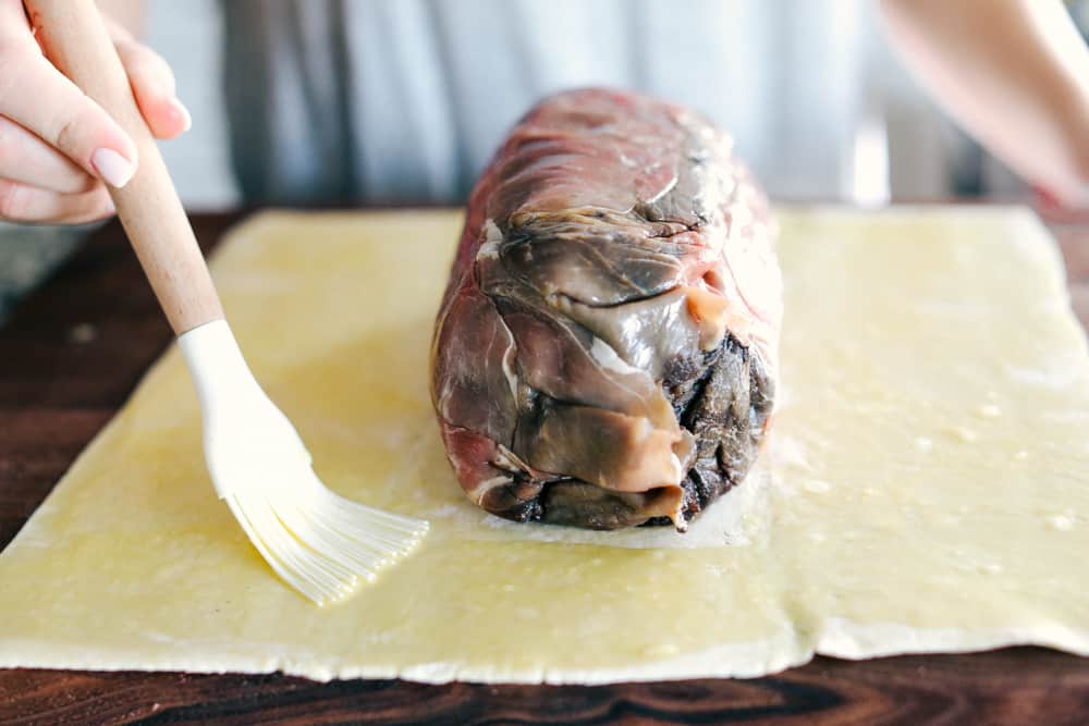 Best Ever Beef Wellington Recipe is a decadent tender and juicy beef tenderloin with a layer of mustard, mushroom sauce and prosciutto wrapped in a soft, buttery pastry that is cooked until golden brown. A holiday favorite! Side shot of someone brushing butter on to the pastry.