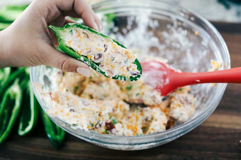 These jalapeรฑoย poppersย are filled with a cream cheese mixture, topped with crumbled bacon pieces, and then baked to perfection. You won't be able to stop at one!ย Jalapeรฑo poppers being stuffed with cream cheese, cheese and bacon mixture.