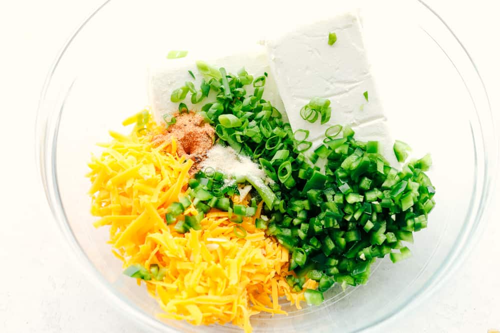 This jalapeno bacon cheese ball is perfectly creamy, rolled in crunchy bacon, and topped off with green onions and salty pecans. It's the perfect holiday appetizer and filled with SO much flavor! Cheese ball ingredients being added to a glass bowl.