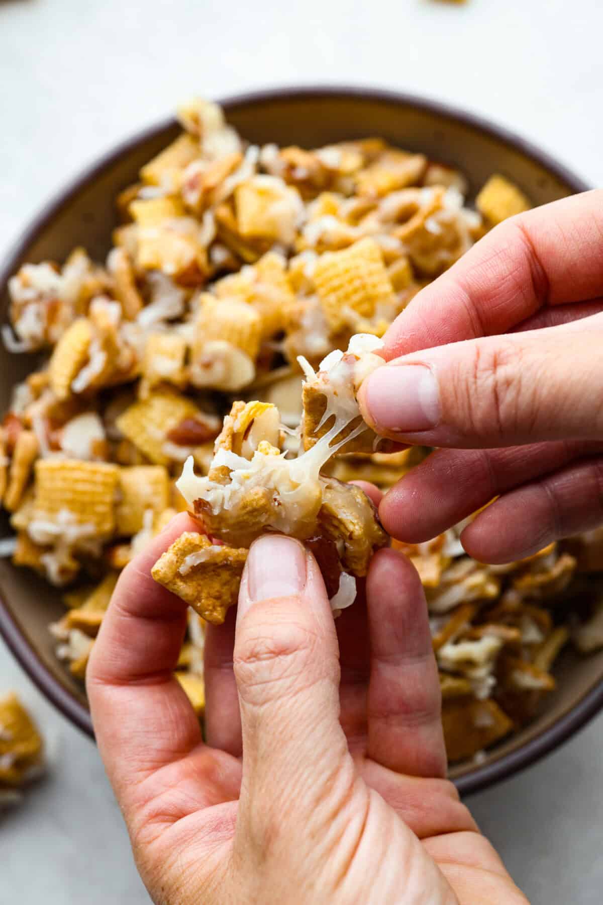 Ooey Gooey Chex Mix is the perfect sweet, salty, and crunchy blend. It has a mixture of cereal, sliced almonds, and shredded coconut, all covered in a soft caramel sugar mixture. This is a must-make for the holiday season! Close up shot of someone stretching a chunk of ooey gooey Chex mix between their fingers.