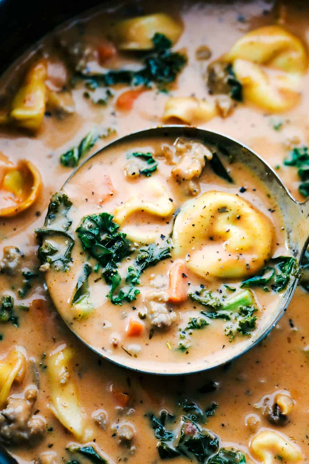 Creamy sausage tortellini soup is thick and creamy with chunks of cooked pork sausage, carrots, buttery kale and three cheese tortellini pasta all cooked in a smooth tomato sauce. This soup will become an instant favorite! Closeup of tortellini soup in a ladle.