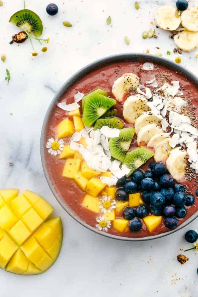 The Best Acai Bowls (Three Ways!) is a fruit filled bowl full of acai berries blended together with a juice topped with granola, fresh fruits, crunchy nuts and a variety of seeds all drizzled with a fun topping. A perfect way to start your day or keep it going! Tropical Mango acai bowl with fruit toppings and a fruit ingredients on the side.