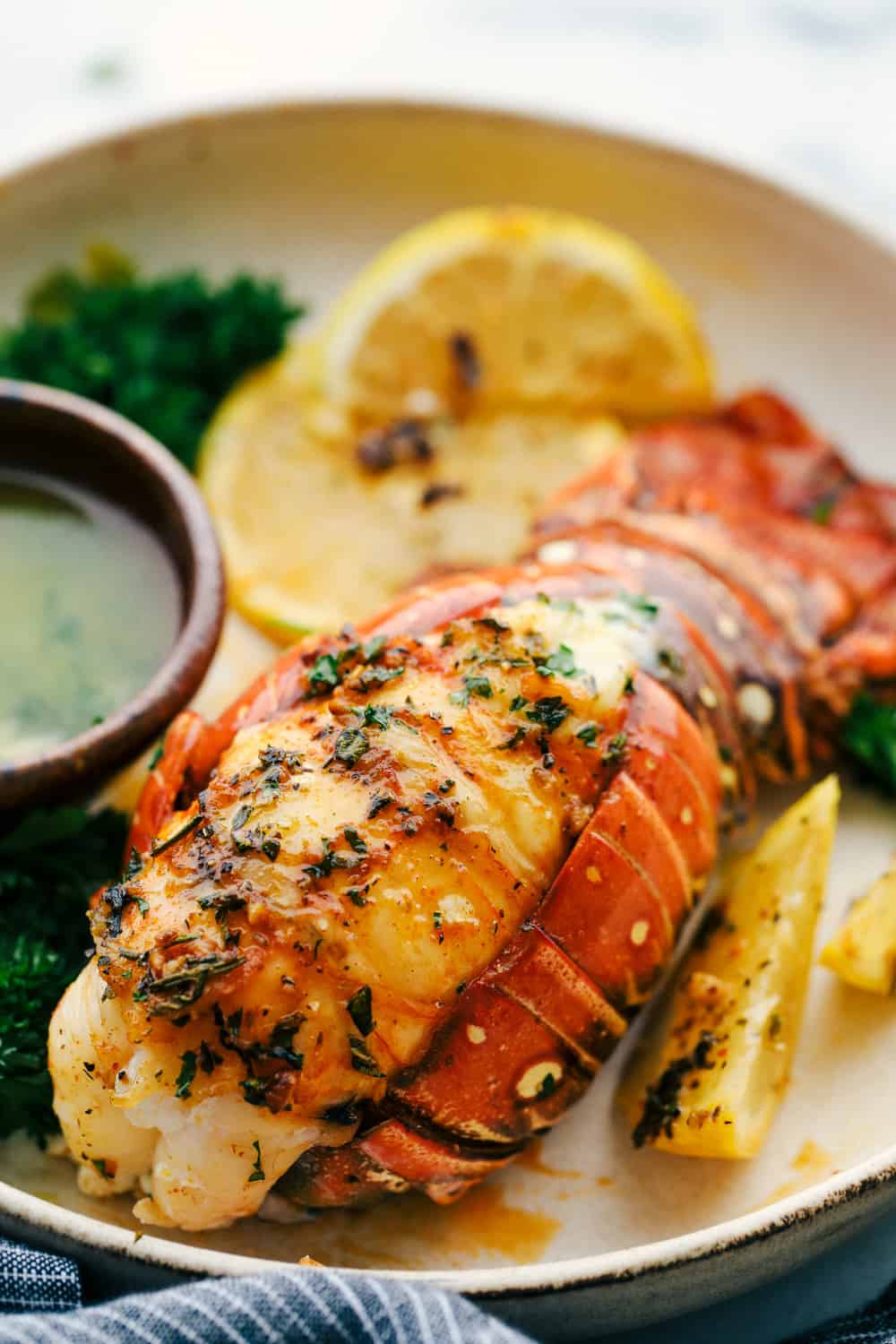 The Best Lobster Tail Recipe Ever is a decadent dinner made with large lobster tails smothered with a buttery garlic herb sauce then broiled under high heat making these lobster tails tender and juicy. The ultimate indulgence! A lobster tail butterflied, and served on a plate with a buttery garlic herb sauce with lemon slices.