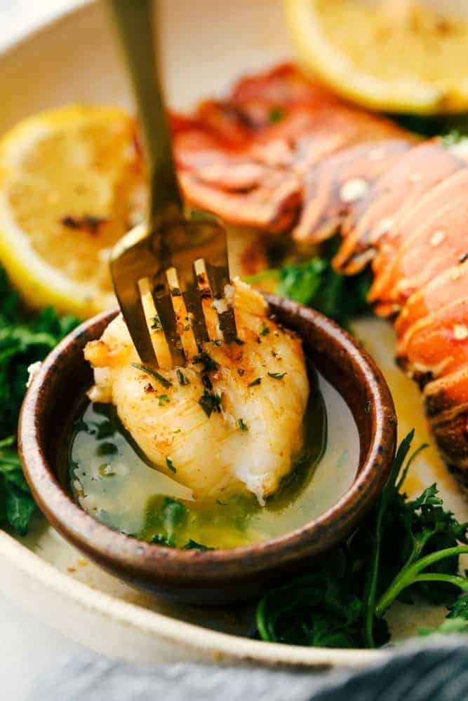 The Best Lobster Tail Recipe Ever is a decadent dinner made with large lobster tails smothered with a buttery garlic herb sauce then broiled under high heat making these lobster tails tender and juicy. The ultimate indulgence! Lobster meat on a fork being dipped in a buttery herb garlic sauce.