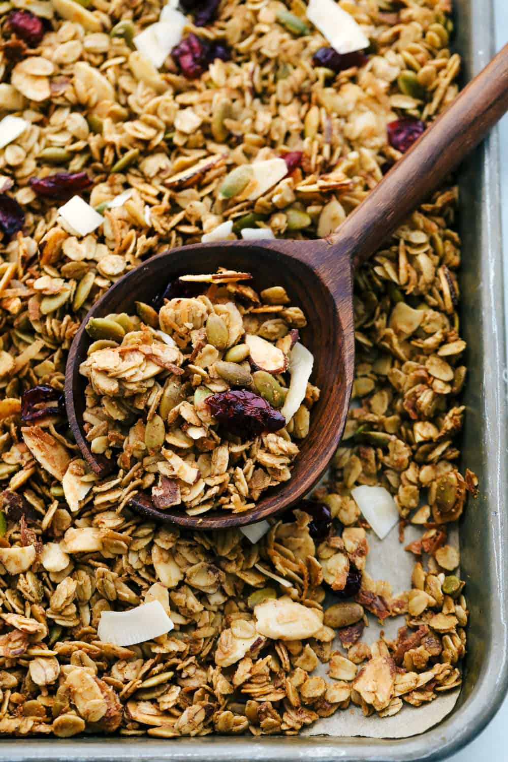 Insanely Good Homemade Granola is a sweet and savory granola with oats, nuts, seeds, dried fruit mixed together with a cinnamon seasoning and combined with a peanut butter and maple syrup poured over top, then baked to perfection. This is a MUST make granola in 20 minutes! Homemade baked granola on a sheet pan with a large wooden spoon scooping it up.