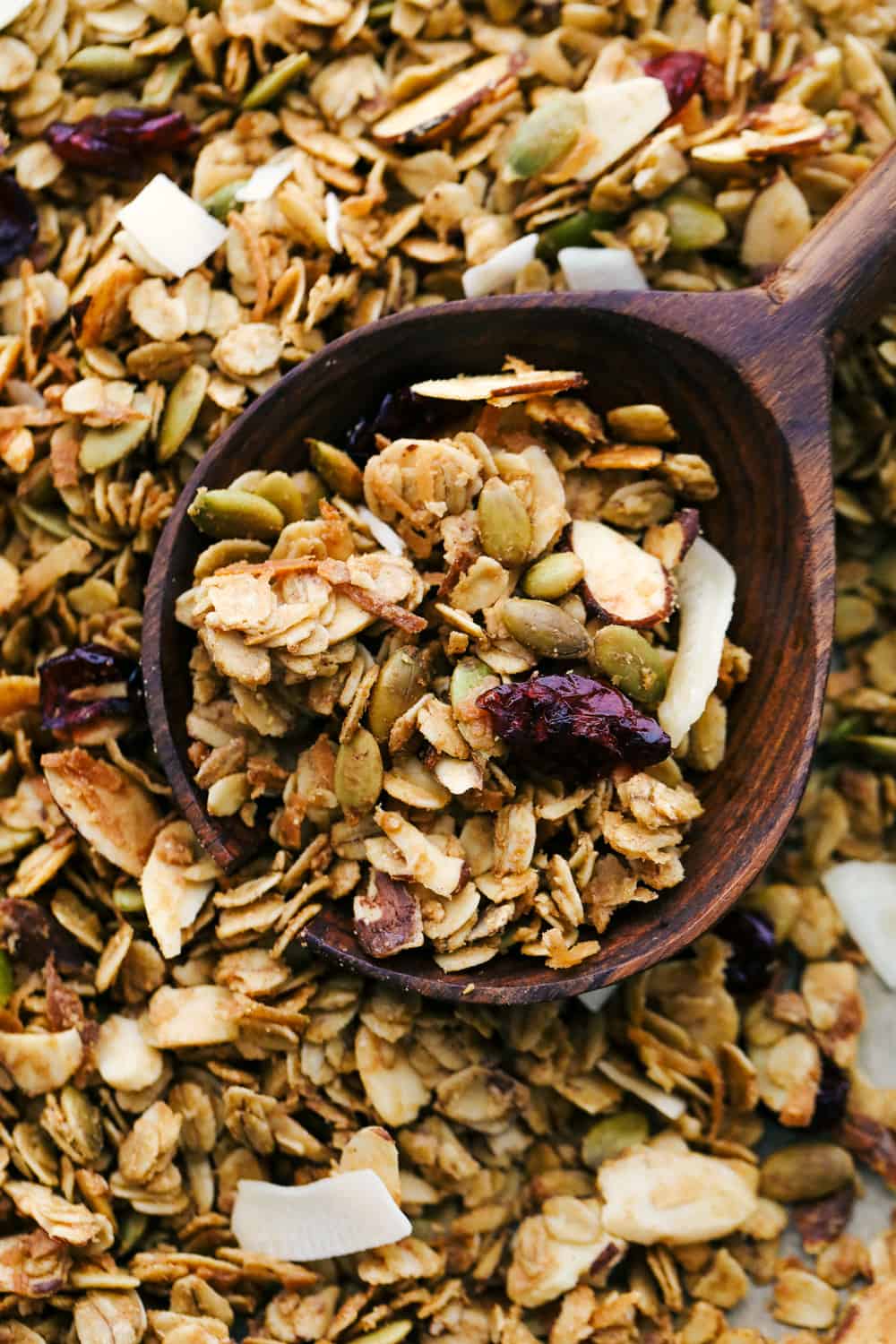 Insanely Good Homemade Granola is a sweet and savory granola with oats, nuts, seeds, dried fruit mixed together with a cinnamon seasoning and combined with a peanut butter and maple syrup poured over top, then baked to perfection. This is a MUST make granola in 20 minutes! Homemade granola up close photo with it baked on a sheet pan and a wooden spoon.