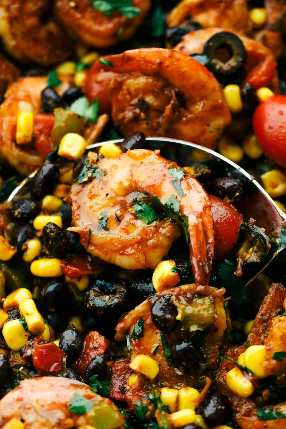 Mexican Shrimp Skillet is full of thick, plump shrimp with black beans, goldenย yellowย corn, ripe cherry tomatoes, chopped bell peppers, black olives mixedย together in myย famous tacoย seasoning all inย sautรฉd in one pan. All under 20 minutes! Mexican Shrimp Skillet with an up close photo with a spoon and shrimp on it.