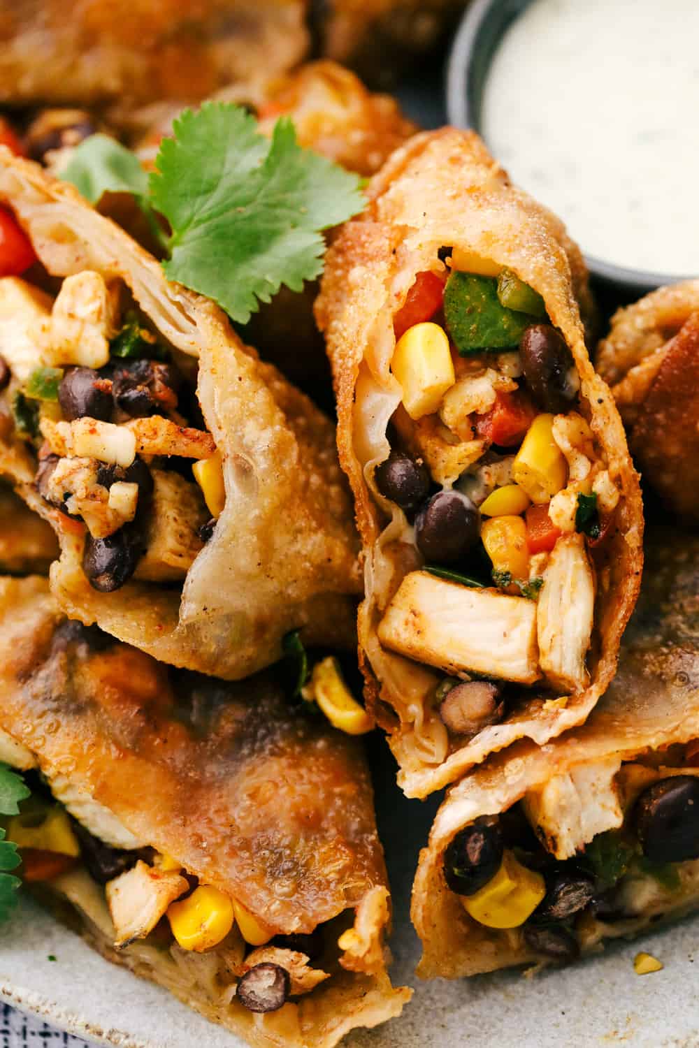 These are the BEST Southwest chicken egg rolls! It's aย colorfulย combination of corn, black beans, fresh veggies, shredded cheese, and chicken. This tasty mixture is flavored in savory southwestย seasonings then fried in a crispy egg roll wrapper. These won't last long at your next party! Southwest chicken egg rolls cut in half with the cilantro ranch dressing on the side.