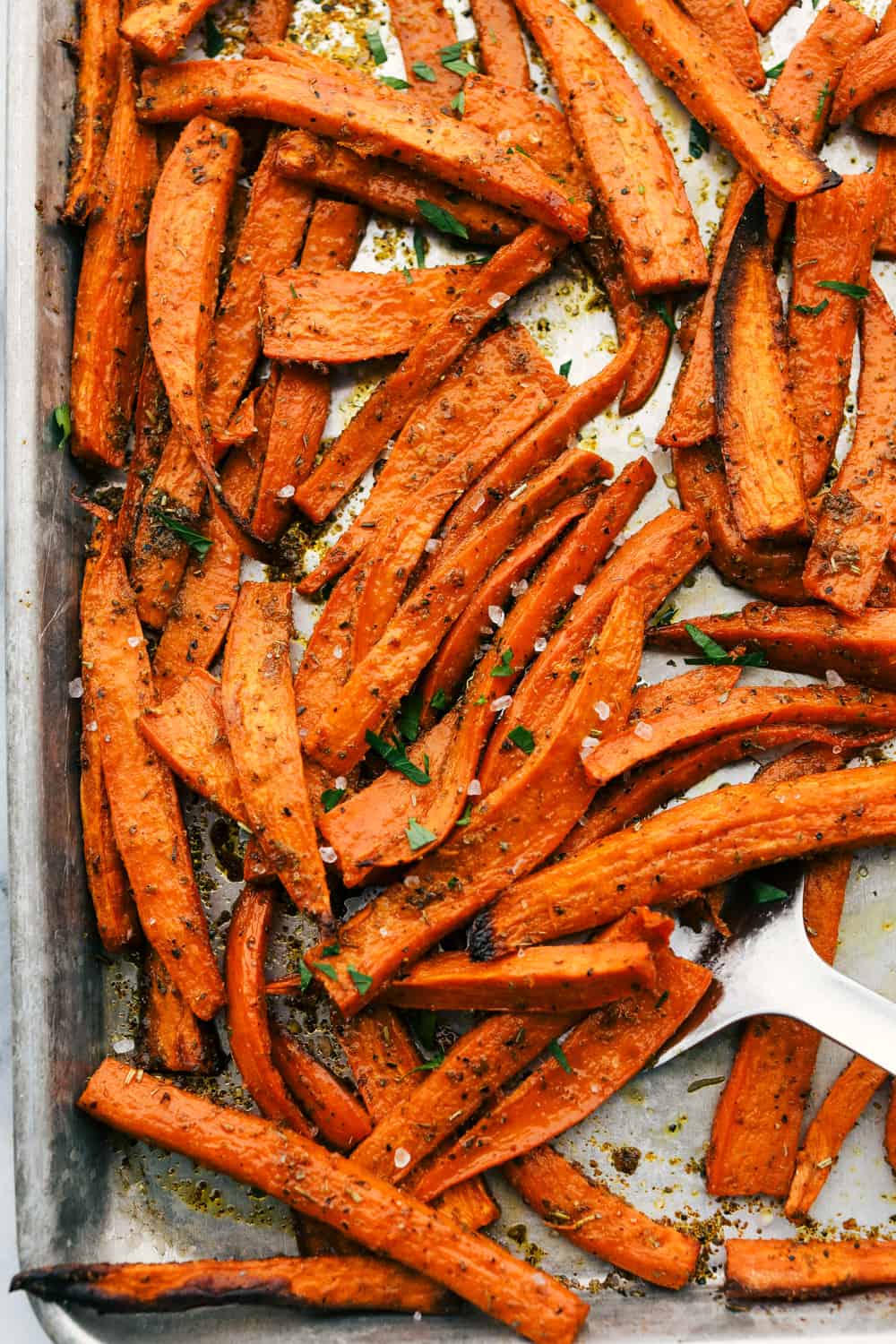 Crispy Garlic Sweet Potato Fries are thick cut sweet potatoes baked in olive oil and then seasoned with garlic powder and Italian seasoning for a crispy outside and a tender inside. The perfect side dish or snack! Crispy garlic sweet potato fries on a pan baked with a spatula scooping them out.