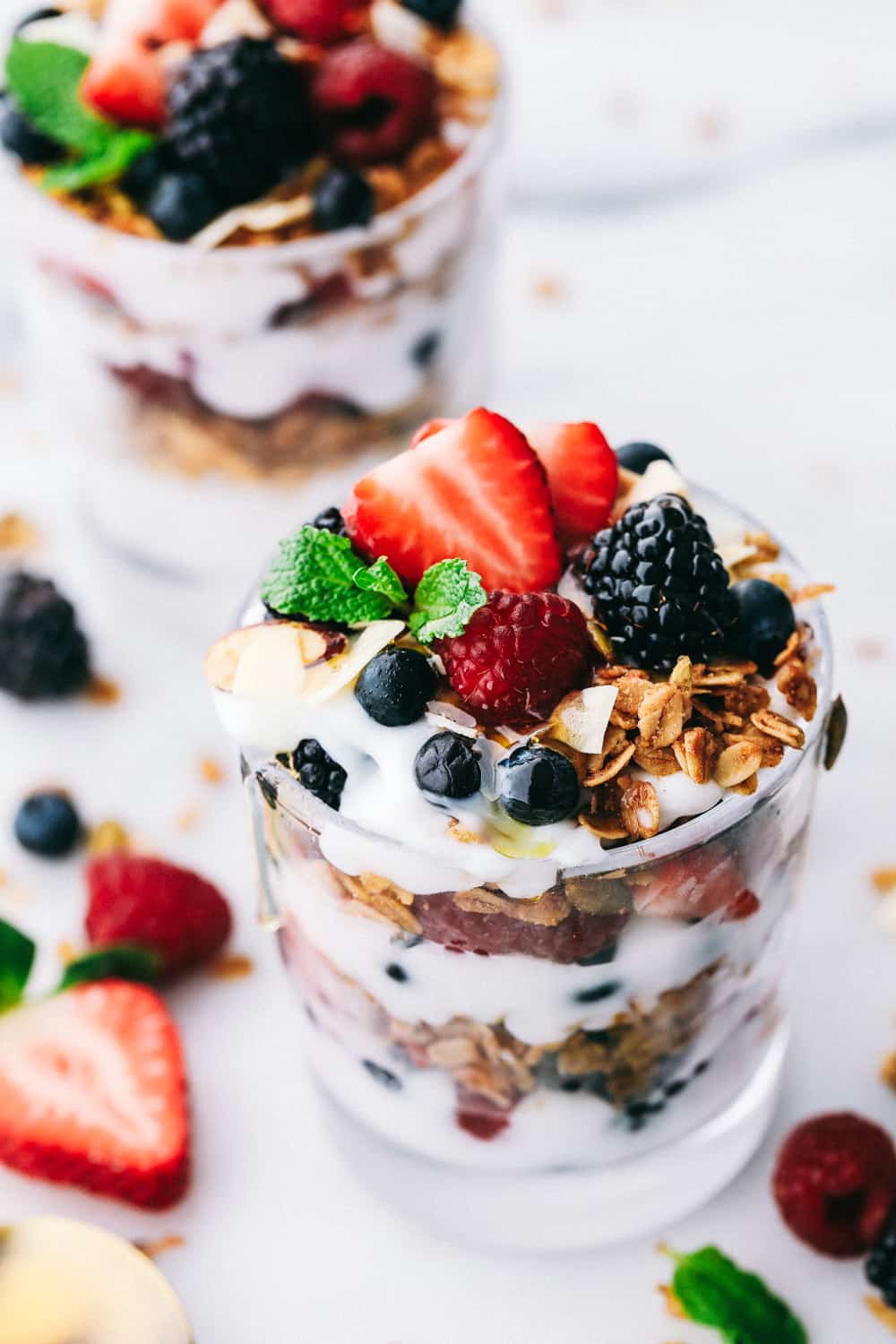 Yogurt Parfait recipe is a simple breakfast or snack that starts with a thick and creamy yogurt layered with homemade granola and fresh berries then topped with more fresh berries and a honey drizzle. A delicious breakfast ready in only a few minutes! yogurt parfaits layered in glass cups garnished with mint and berries spread around the glass cups for display.
