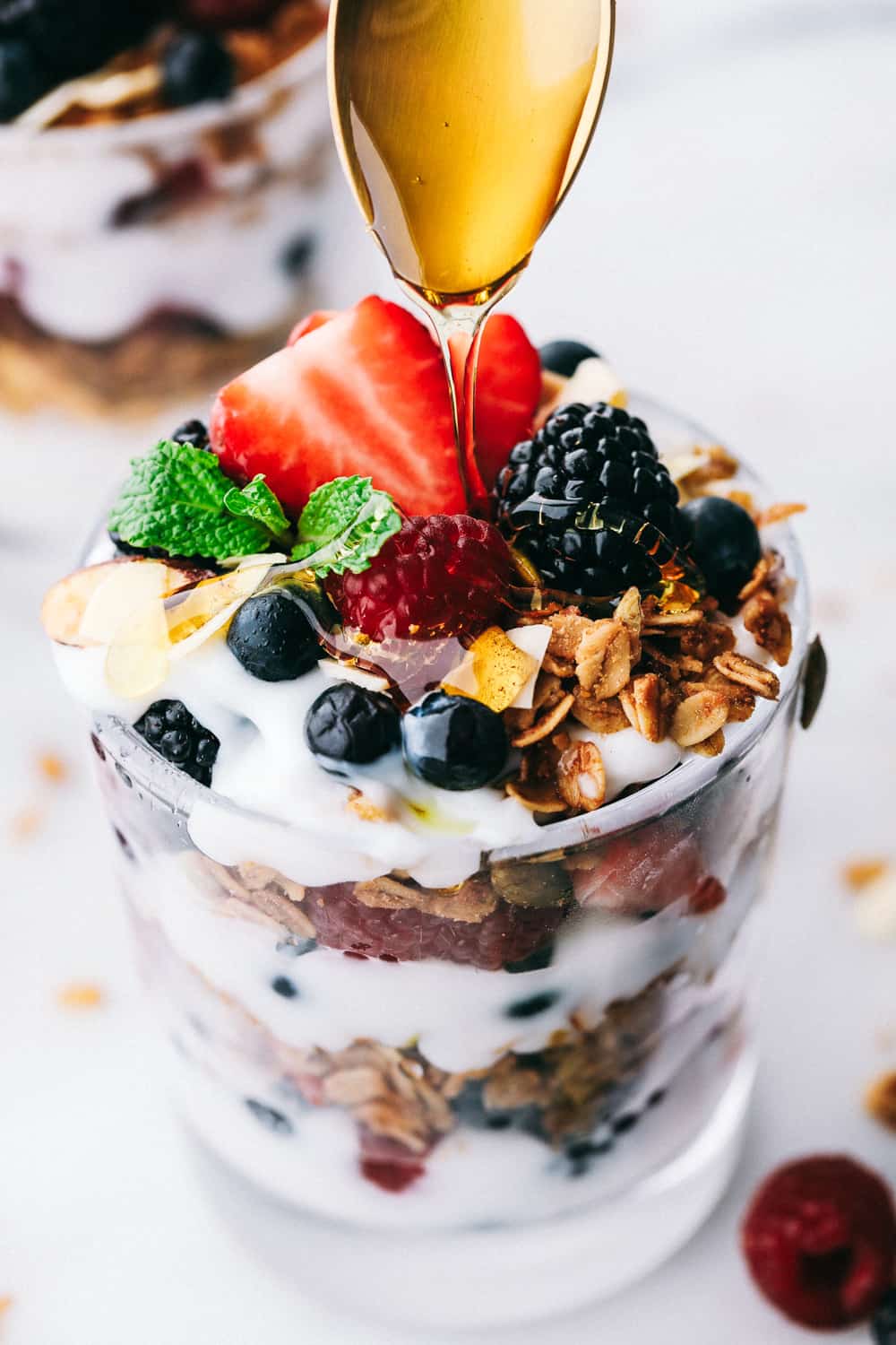Yogurt Parfait recipe is a simple breakfast or snack that starts with a thick and creamy yogurt layered with homemade granola and fresh berries then topped with more fresh berries and a honey drizzle. A delicious breakfast ready in only a few minutes! yogurt parfait in a glass cup with honey being drizzled on by a spoon.
