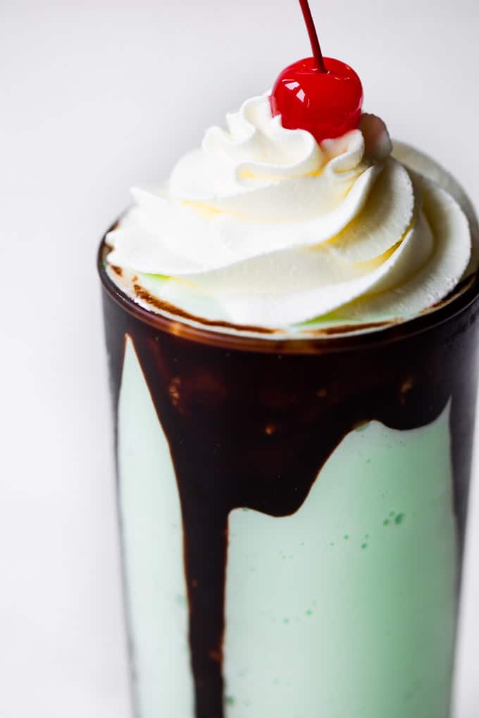 Shamrock Shake is a minty-vanilla ice cream shake is such a fun treat for all ages! Skip the line and make your own at home! Shamrock shake with chocolate syrup dripping down the sides with whip cream topping and a cherry on top.
