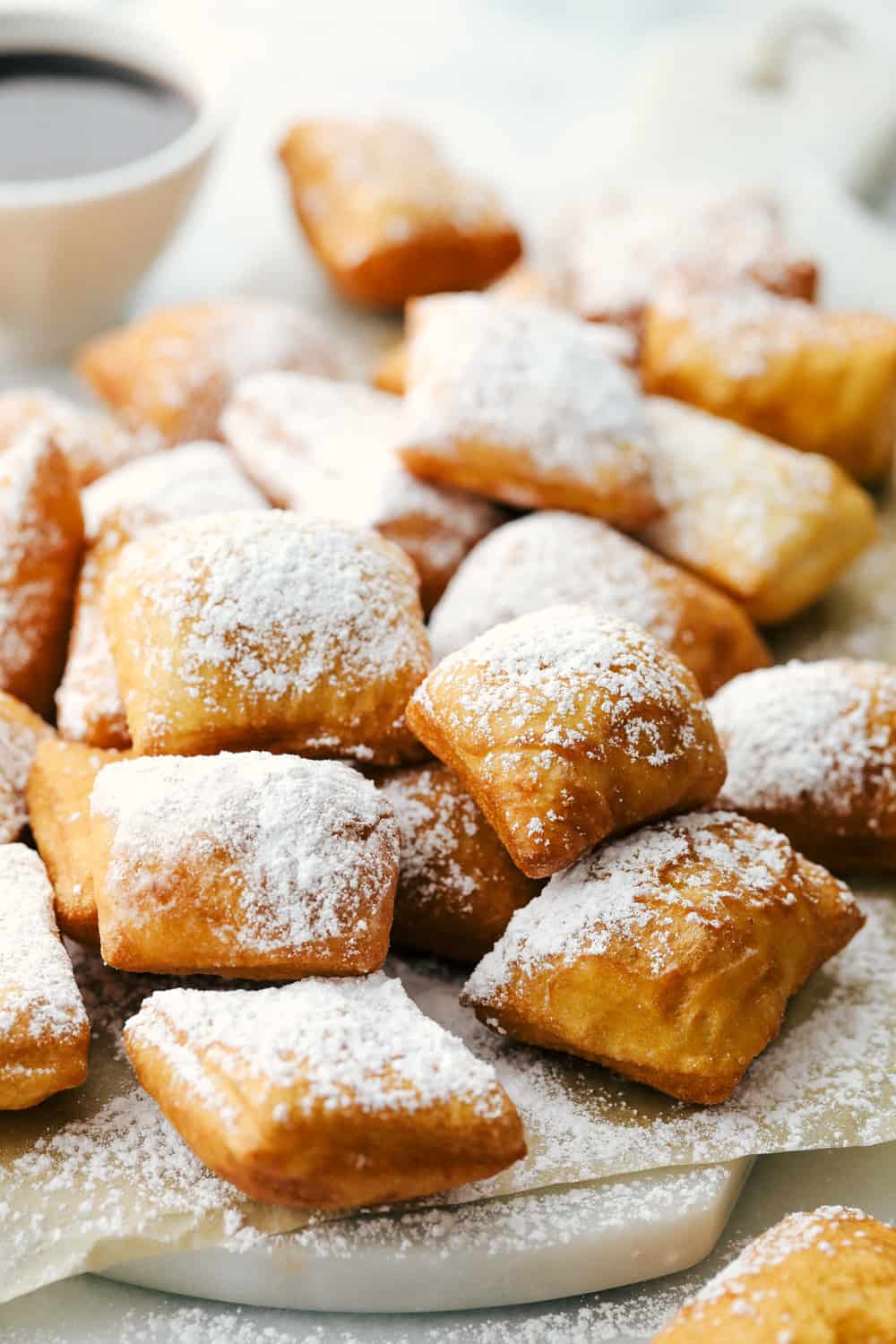 Best Beignets Recipe is a light, fluffyย Frenchย doughnut that are cut into squares and puffed up when fried in oil and then end by sprinkling with powdered sugar or dipped inย chocolate syrup. The best pillowy-soft and fluffy treat to enjoy!ย Several beignets on a cutting board sprinkled with powder sugar and chocolate syrup on the side.