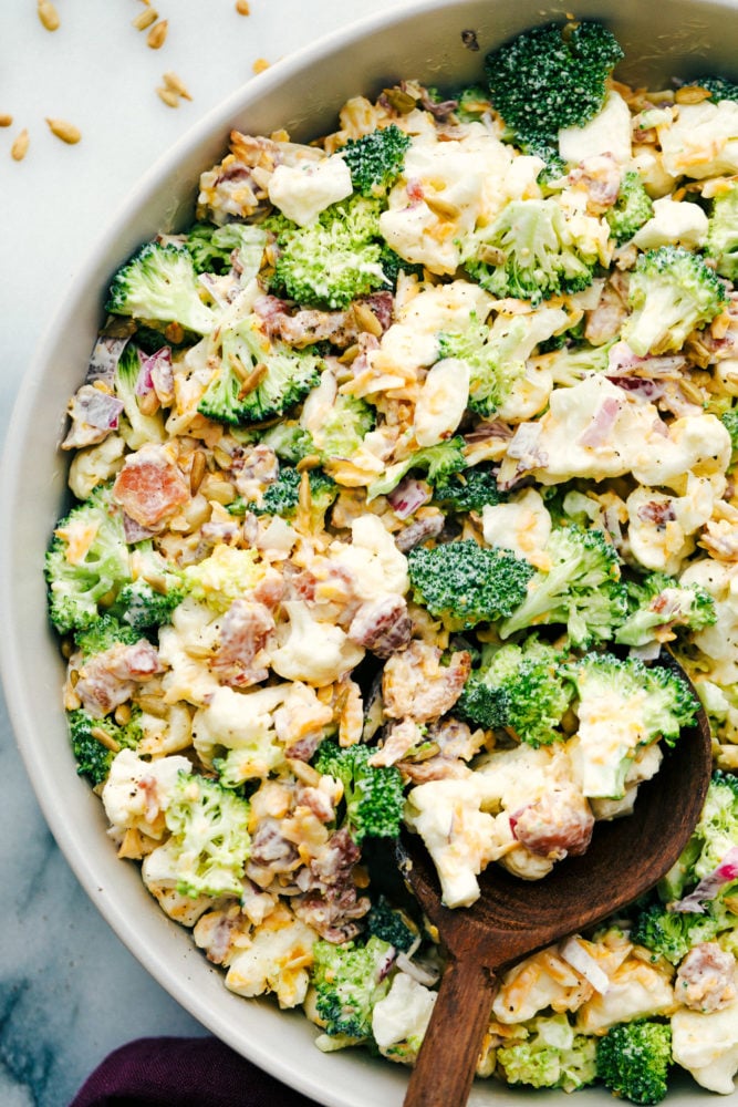 Cauliflower broccoli salad is filled with fresh florets and red onions, bacon pieces with chunks of cheese making this the perfect side salad. Cauliflower broccoli salad mixed together in a bowl with a wooden spoon.