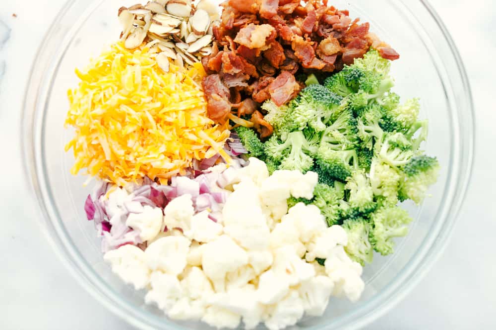 Cauliflower broccoli salad is filled with fresh florets and red onions, bacon pieces with chunks of cheese making this the perfect side salad. Shredded cheese, sliced almonds, bacon chopped up, broccoli and cauliflower florets and red onion chopped up separated in a glass bowl.