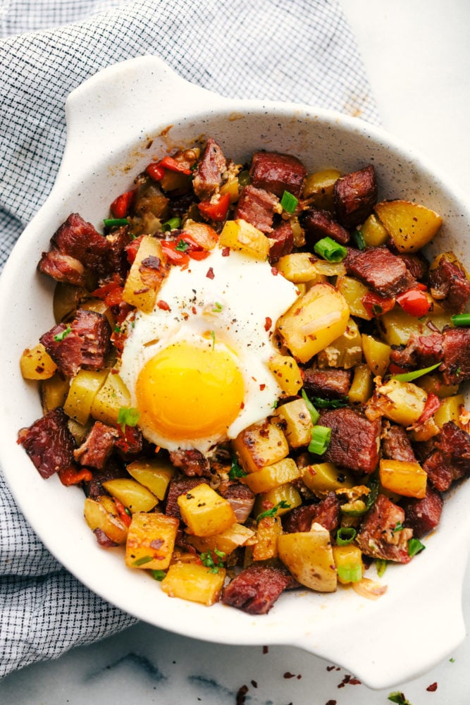 This corned beef hash breakfast is filled with chopped crispy potatoes and bell peppers with corned beef mixed together and a large sunny side up egg to complete the meal. Breakfast just got better with this hash! Corned beef hash with an egg on top in a white skillet.