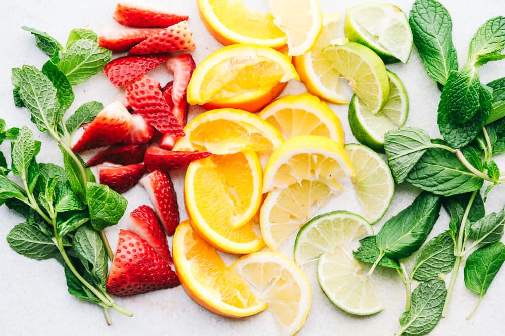 Detox Water is a naturally flavored water mixed with sliced oranges,ย citrus filled lemons and limes, fresh strawberries and garnished with mint making this anย irresistibleย water. Only 5 Minutes to make!ย Photo filled with a mint, sliced strawberries, quartered oranges, lemons and limes with another side of mint on a table.