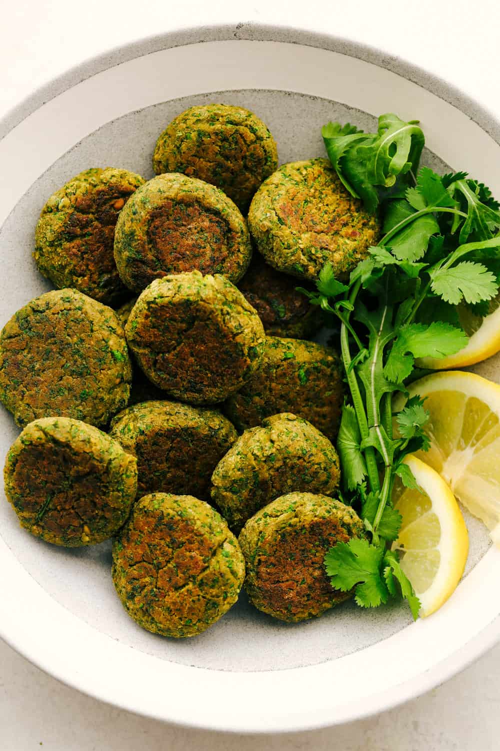 Falafel is a round bite sized mixture of garbanzo beans, seasonings and a hint of spice from a jalapeño all cooked and ready to add to any wrap or salad. A healthy little bite! Falafel on a plate with lemon as a garnish and cilantro.