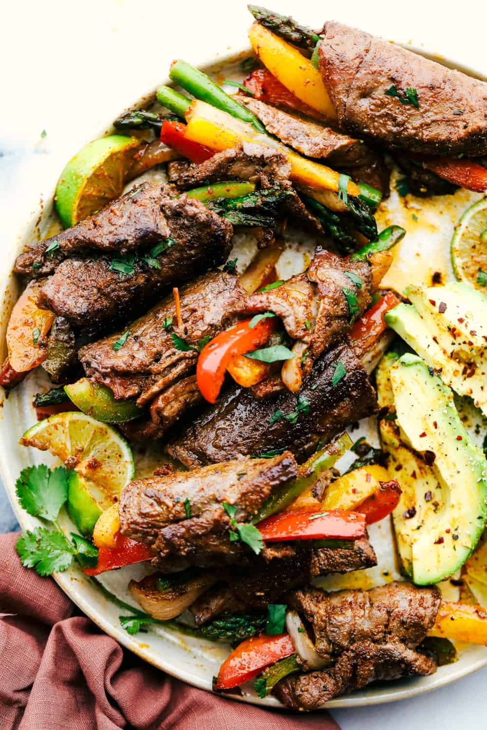 Steak fajita roll ups are fun and festive for the whole family and look extra special sautéed with bright colored bell peppers and fresh asparagus seasoned with my homemade fajita seasoning then rolled up in the flank steak. This makes for a light and delicious dinner! Bell peppers and asparagus wrapped up in flank steak with avocados on a plate.