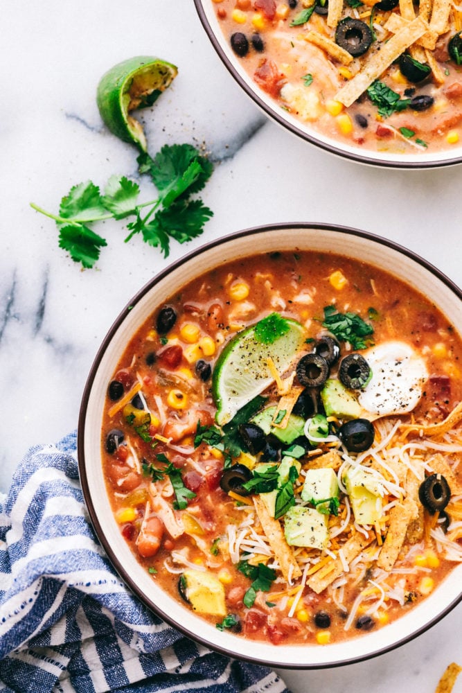 You are going to fall in love with this quick and easy 8 Can Chicken Taco Soup recipe! It is the perfect dinner when there is no time to run to the grocery store but the family needs dinner, like, NOW. Grab these ingredients right out of your pantry and whip up a delicious and flavorful soup! Two bowls of chicken taco soup garnished with sliced olives, limes and a dollop of sour cream.