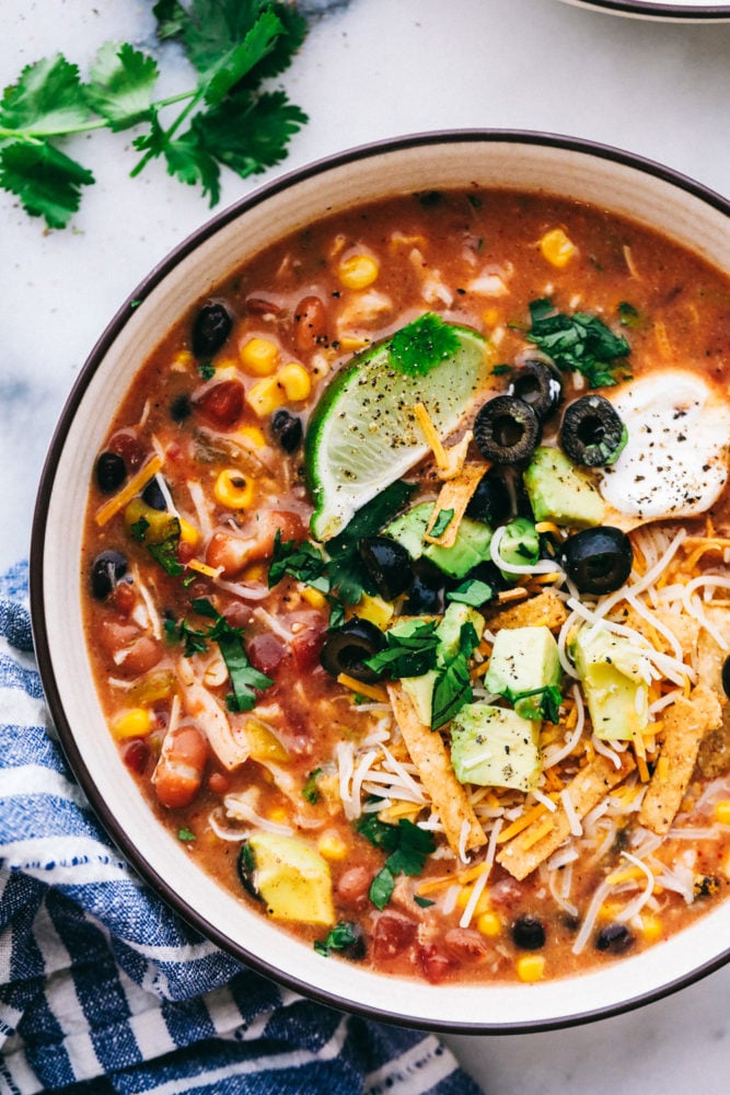 You are going to fall in love with this quick and easy 8 Can Chicken Taco Soup recipe! It is the perfect dinner when there is no time to run to the grocery store but the family needs dinner, like, NOW. Grab these ingredients right out of your pantry and whip up a delicious and flavorful soup! A bowl of 8 can chicken taco soup garnished with lime wedges, sour cream, sliced olives, avocado and tortilla strips.