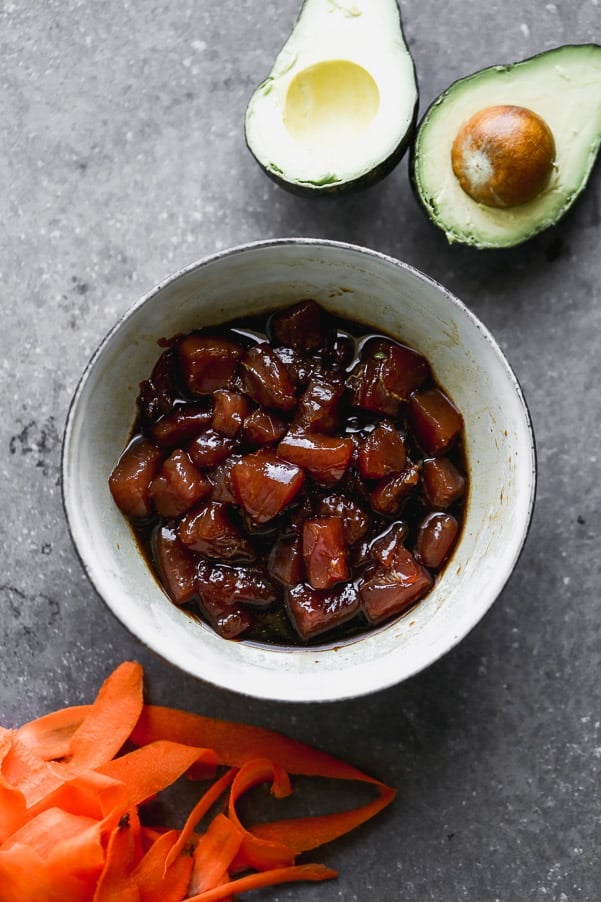 This easy Poke Bowl Recipe ย is packed with sushi-grade ahi tuna seasoned with soy, honey, and plenty of sesame. It's served with sticky brown rice, tons of veggies and these easiest spicy mayo on the planet.ย Ahi tuna being marinated with an avocado cut in half and shredded carrots on the side.