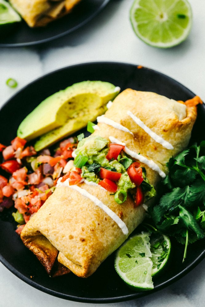 Try these chimichangas either baked or pan fried with tender chicken, stuffed Mexican filling all wrapped in a soft but crispy crust on the outside. These will become a family favorite! Chicken chimichanga on a black plate with an avocado sliced and limes slices on the side.