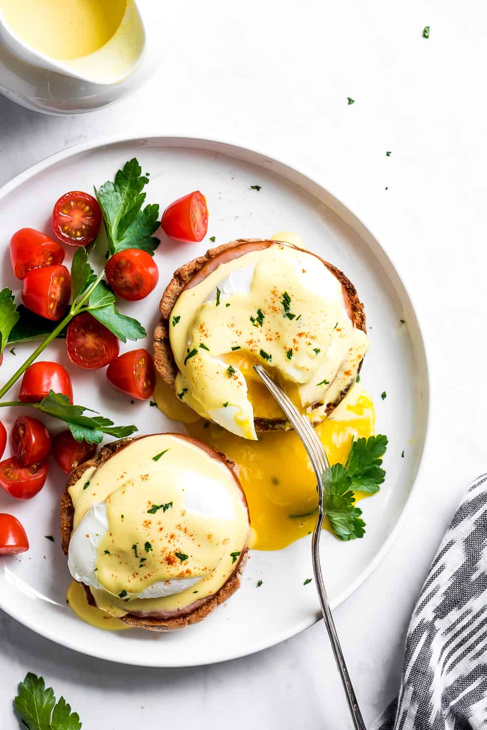 Eggs Benedict is a breakfast favorite! An English muffin is topped with Canadian bacon, a poached egg, and then drizzled with buttery hollandaise sauce. Your next brunch needs this! Top view of cutting into a poached egg on an English muffin.