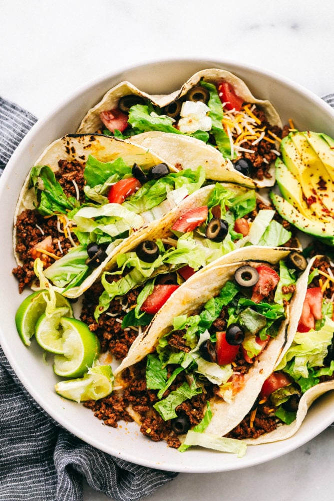 Ground beef tacos are a quick and easy dinner that your whole family will love! Fill a taco with this tender and juicy beef that will become the best weeknight dinner.ย Five beef tacos on a plate loaded with shredded lettuce, sliced black olives, shredded cheese, tomato slices with lime slices and an avocado sliced on the side.