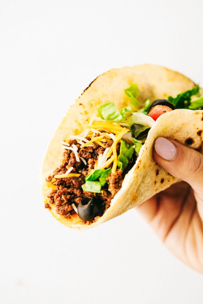 Ground beef tacos are a quick and easy dinner that your whole family will love! Fill a taco with this tender and juicy beef that will become the best weeknight dinner.ย A beef taco with olives, shredded lettuce and shredded cheese being held up with the recipe critic hand.