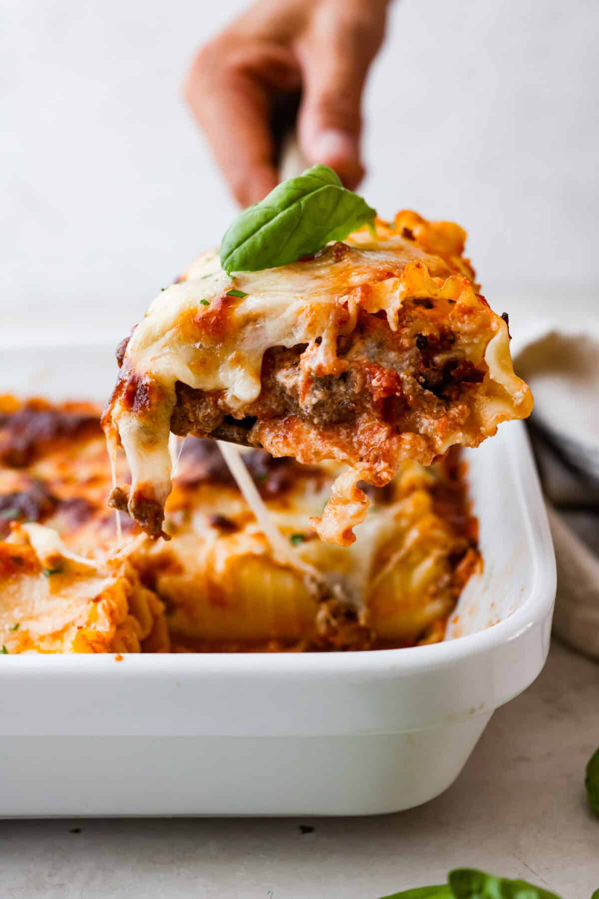 These Lasagna roll-ups have all the flavors of a classic lasagna wrapped up in a fun lasagna twist! This recipe is an easier approach to this classic Italian comfort food; seasoned beef, sweet marinara sauce, and melty, creamy cheese wrapped in a perfectly al dente noodle. Side shot of someone lifting a lasagna roll up with a basil leaf on top, out of the baking dish.