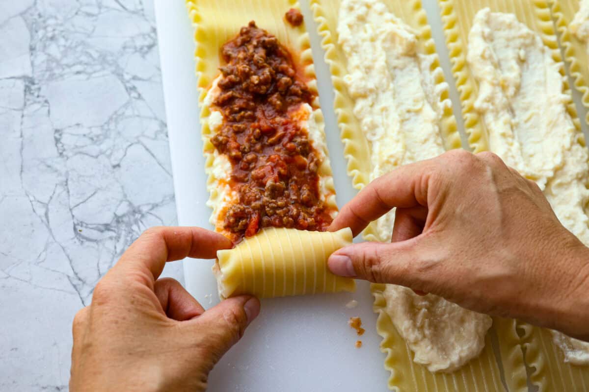 These Lasagna roll-ups have all the flavors of a classic lasagna wrapped up in a fun lasagna twist! This recipe is an easier approach to this classic Italian comfort food; seasoned beef, sweet marinara sauce, and melty, creamy cheese wrapped in a perfectly al dente noodle. Overhead shot of someone rolling up the lasagna noodle with cheese and sauce spread on top.