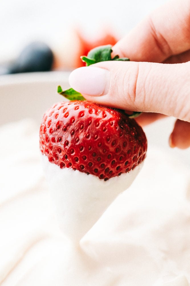 Marshmallow cream cheese fruit dip is one of the best dips to enjoy year round. Fluffy fruit dip is sweet and creamy that is served with all your favorite fresh fruits!ย A red strawberry being dipped in marshmallow cream cheese fruit dip.