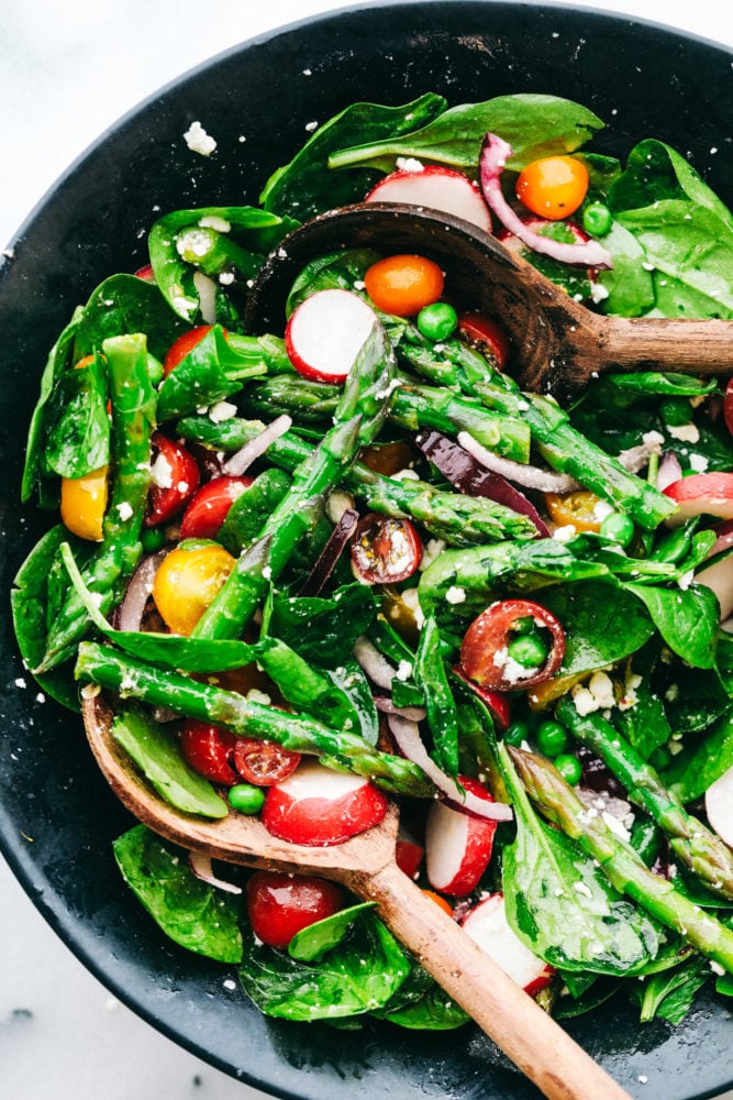 This fresh asparagus salad is one of my favorites and I can't wait to show you how to make it. With lemon vinaigrette dressing drizzled on top this salad has a light and flavorful crisp crunch in every bite. Asparagus salad being tossed together in a black bowl with two wooden spoons.